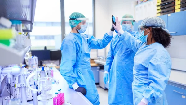 Ba nhà khoa học mặc đầy đủ trang phục bảo hộ cá nhân (PPE), bao gồm áo choàng, khẩu trang và tấm che mặt, đang vỗ tay chúc mừng nhau trong phòng thí nghiệm.